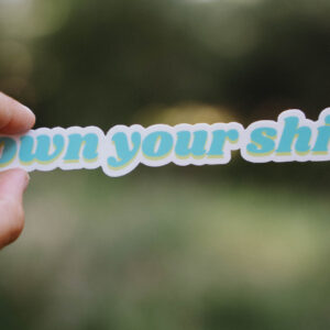 Sticker that says "own your shit" held up in front of a green background