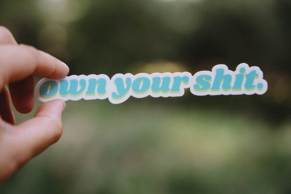 Sticker that says "own your shit" held up in front of a green background