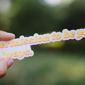 Sticker that says "Permission to be you. Always." held up in front of a green background