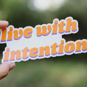 Sticker that says "live with intention" held up in front of a green background