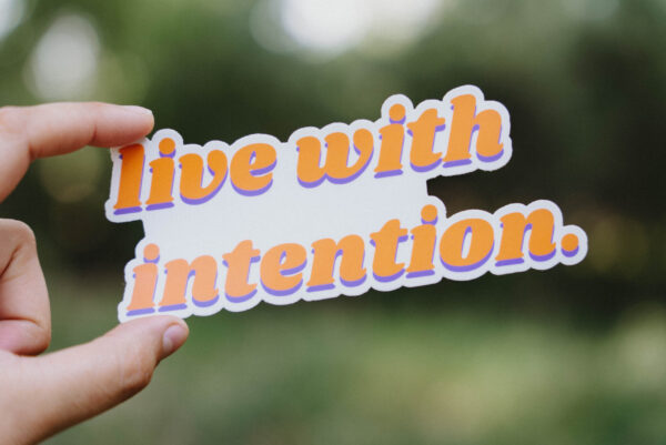 Sticker that says "live with intention" held up in front of a green background